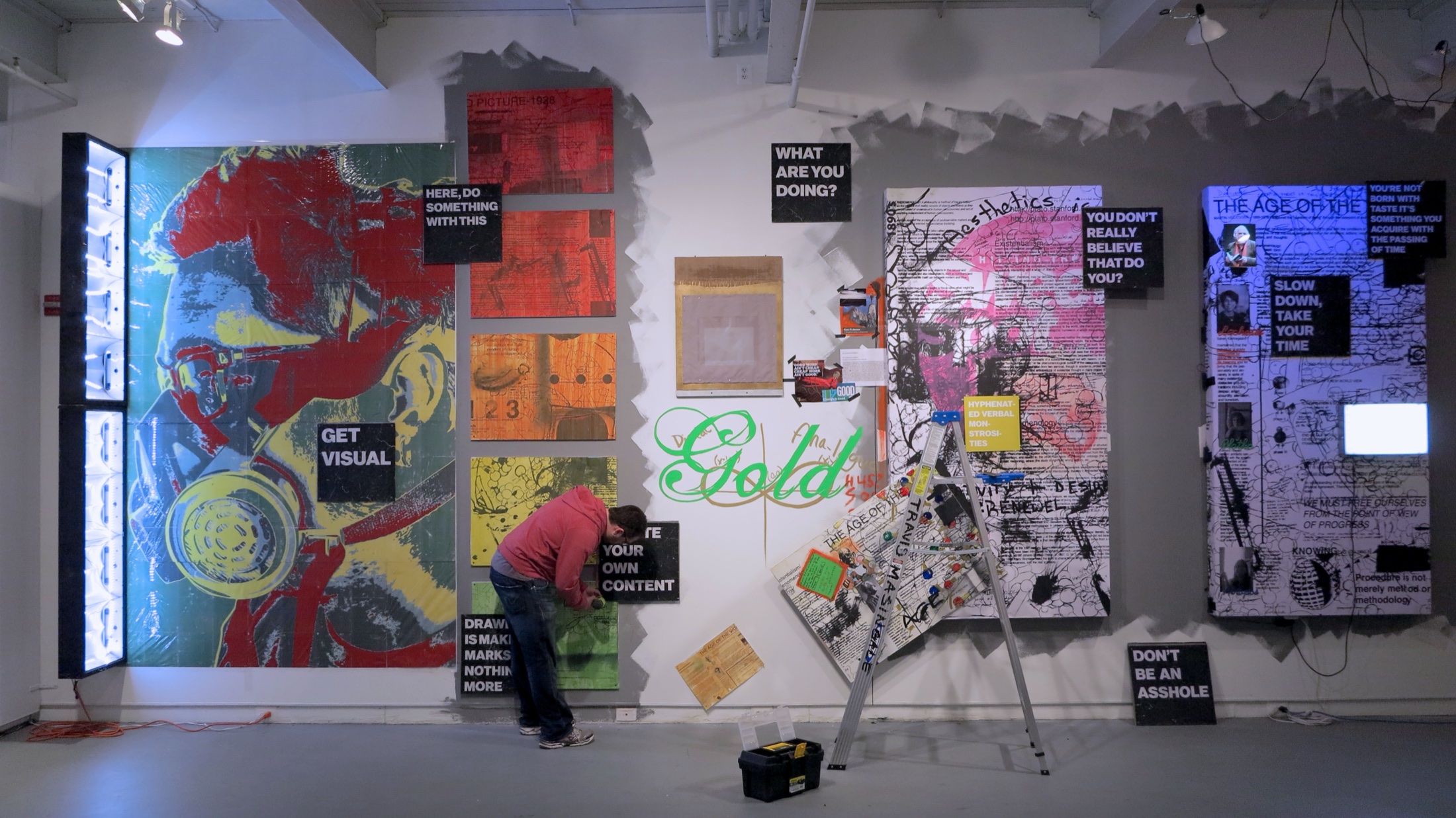A person in a red hoodie works at the exhibition wall beside a ladder, toolbox, loose panels, and a large arrangement of images and text.