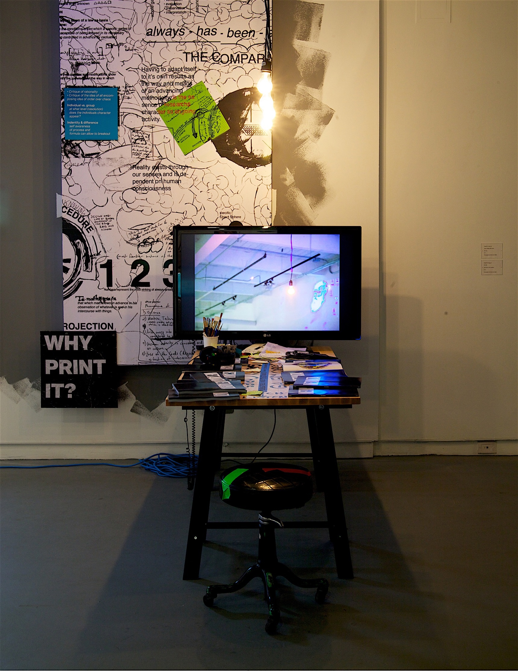 Installation view with a worktable, monitor, studio stool, hanging bulb, and a layered wall composition behind them.
