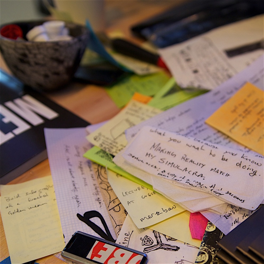 A worktable is covered with handwritten cards, scraps, diagrams, and sticky notes.